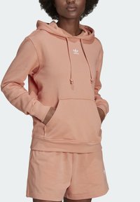 Rosa bomullshoodie med känguruficka, justerbara snören och en liten vit logotyp på bröstet, ihop med matchande shorts.