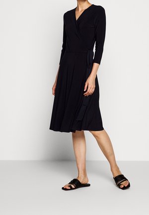 Woman wearing a black wrap dress with three-quarter sleeves and black flat slide sandals, standing against a plain light background.