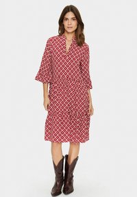 Röd mönstrad klänning med krage, trekvartslånga ärmar och lager av volanger. Bärs med bruna cowboyboots. Bomullstyg, lätt textur.