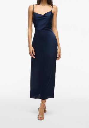 Kvinde iført en ærmeløs midi-kjole i marineblå satin med tynde stropper og guldfarvede åben tå-sandaler med hæl, stående foran en hvid baggrund.