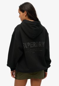 Felpa nera oversize con cappuccio e maniche lunghe. Presenta un grande logo "SUPERDRY" grigio sul retro. Abbinata a una gonna verde oliva.