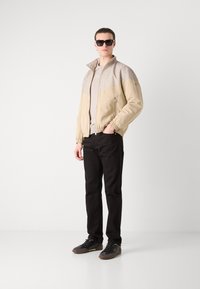 Corta-vento bege com um painel superior texturizado, fecho de correr na frente, punhos elásticos, combinado com calças pretas e sapatos escuros. Óculos de sol usados.