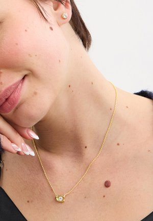 Donna che indossa una collana d'oro con pendente in pietra trasparente e orecchino a bottone, mostra dei nei sulla pelle del collo e della clavicola, mano vicino al viso.