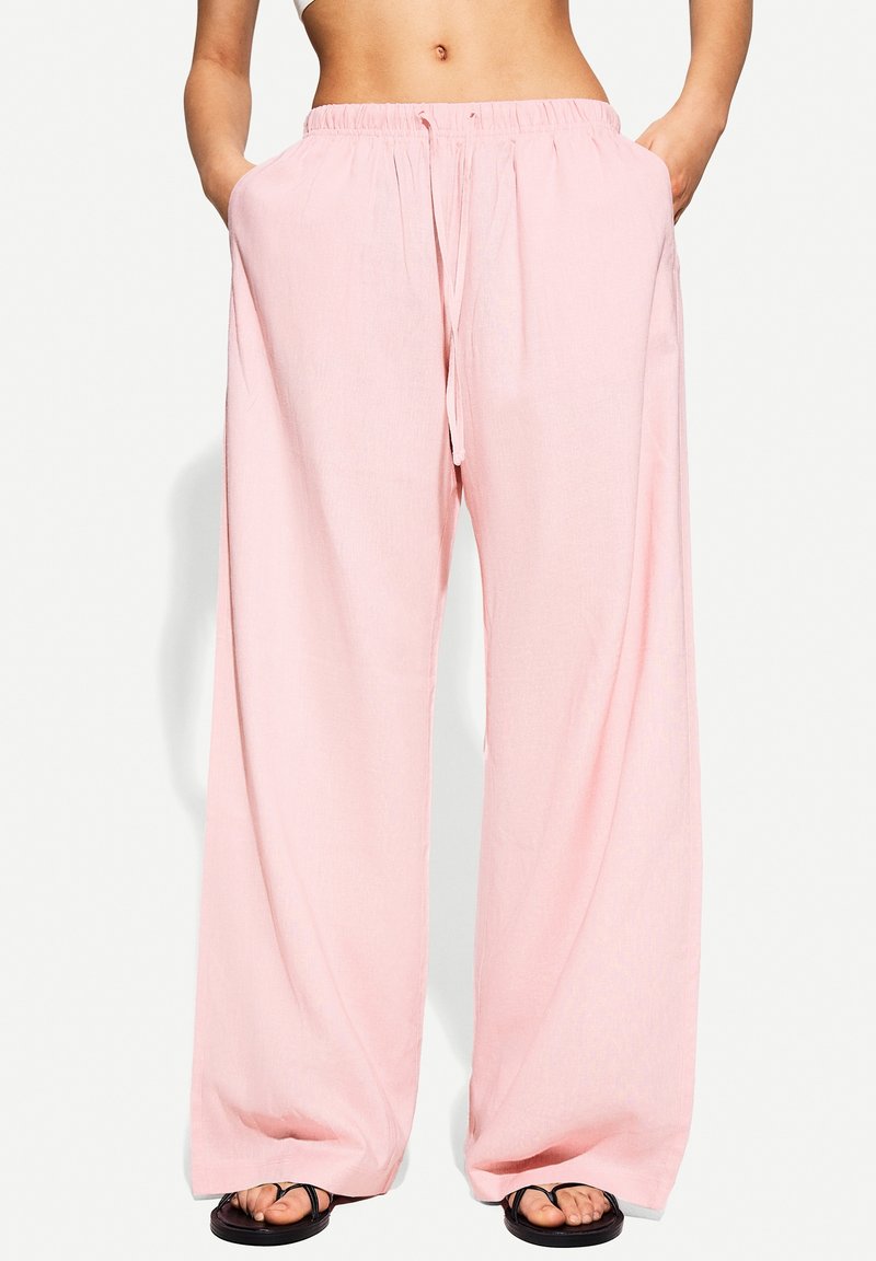 Pantalon ample rose clair avec taille élastique et cordon, porté par une personne avec les mains dans les poches et des sandales noires visibles aux pieds.