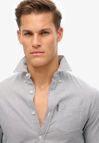 Camicia a bottoni in tessuto a righe, con colletto a contrasto, tasca frontale e un sottile etichetta logata. La combinazione di colori è blu e bianco.