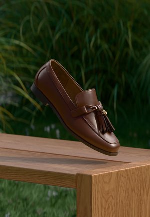 Bruine leren loafer met kwastjes, balancerend op de rand van een houten tafel, met groene bladeren wazig op de achtergrond.