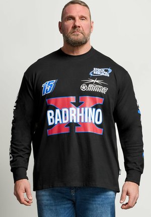 Brodaty mężczyzna z krótkimi włosami, ubrany w czarną koszulę z długim rękawem z logo "BADRHINO" i grafikami w stylu wyścigowym, w połączeniu z niebieskimi dżinsami.