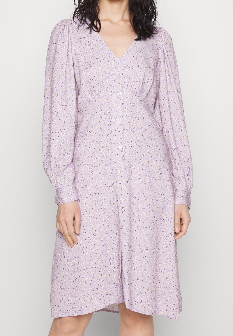 Femme portant une robe violet clair à manches longues, boutonnée, avec de petits motifs floraux et un col en V.