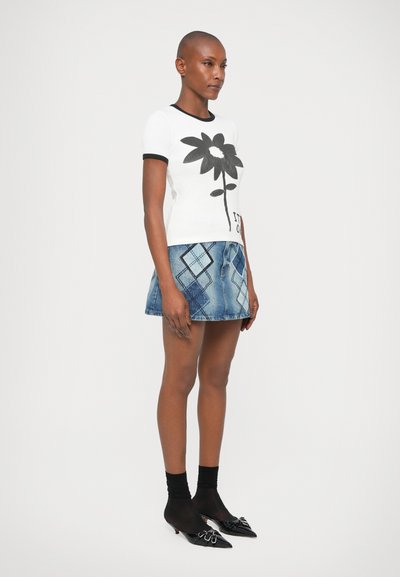 Kvinde med barberet hoved iført hvid T-shirt med stor sort blomst, blå ternet denim mini nederdel, sorte gennemsigtige strømper og sorte spidse sko.