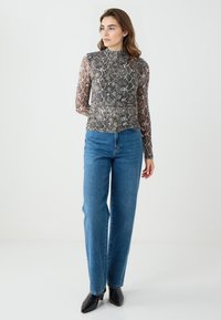 Durchsichtige, langärmelige Bluse mit einem Schlangenmuster in Grautönen und Beige, kombiniert mit hochgeschnittenen blauen Jeans und schwarzen High Heels.