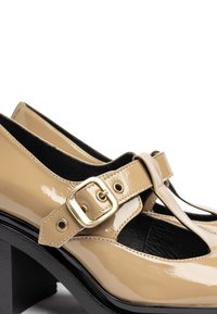 Chaussures en cuir verni beige avec un design à bride T, dotées d'une boucle dorée et d'un large talon noir épais. Doublure intérieure noire texturée.