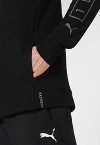 Sudadera con capucha negra, con un bolsillo frontal, puños acanalados y un detalle de etiqueta en el dobladillo. Presenta un logo gris de Puma y texto en la manga.