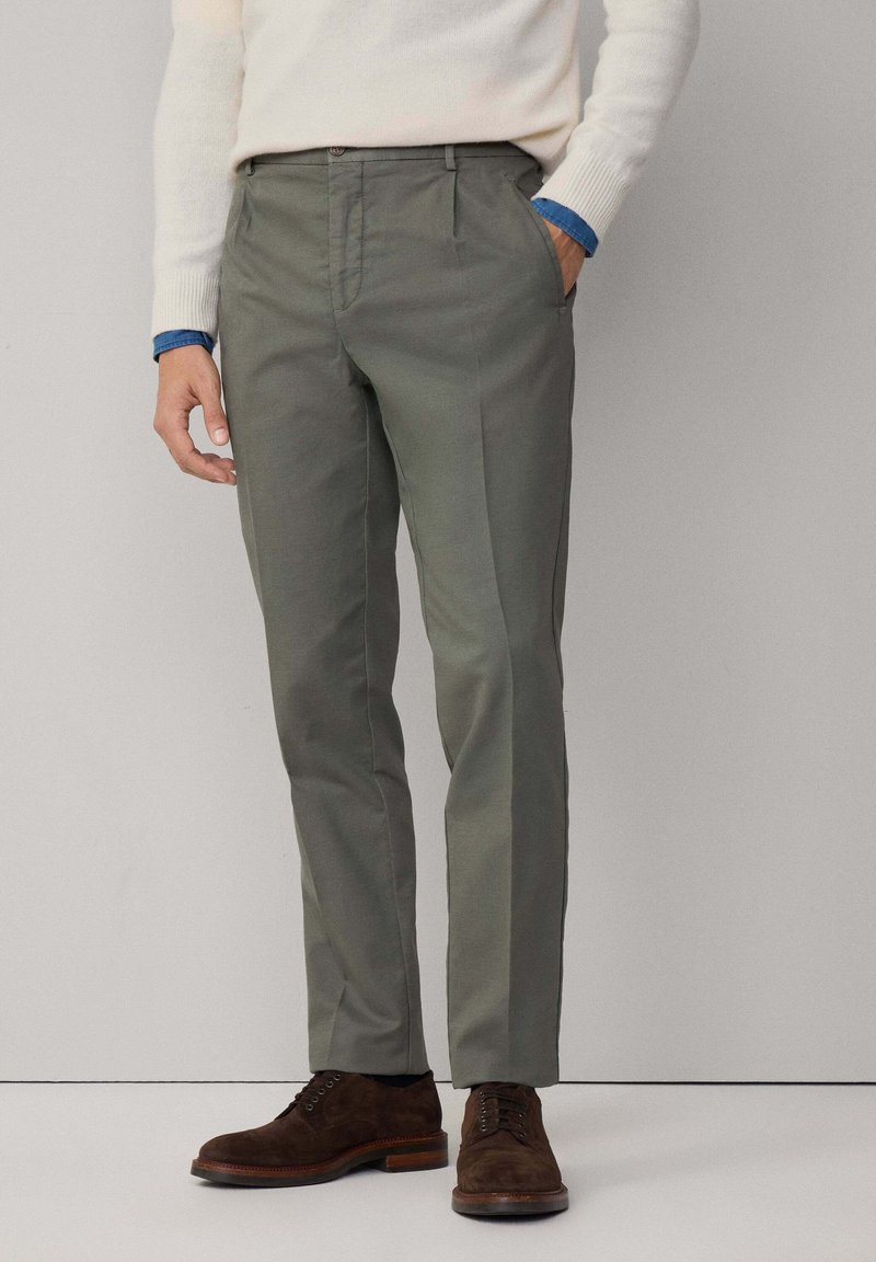 Svetlo šedo-zelené nohavice so slim fit strihom, so splošteným predkom, bočnými vreckami a hladkou textúrou. V páre s tmavohnedými topánkami.