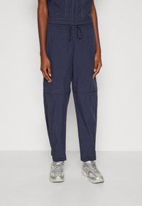 Marineblauwe cargo broek met elastische tailleband en trekkoord. Voorzien van met een rits afgesloten zakken, contrasterende stiksels en een losse, taps toelopende pasvorm.