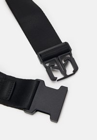 Svart justerbar rem med en plastspänne med sidofäste, visad oöppnad mot en vit bakgrund.