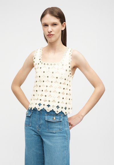 Femme aux cheveux bruns raides portant un haut en dentelle crème sans manches avec des découpes circulaires et un jean bleu taille haute avec poches avant.