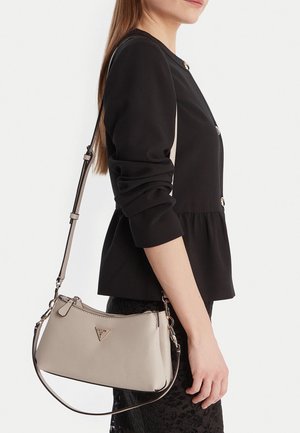 Woman wearing black long-sleeve top and lace skirt, carrying beige crossbody handbag with adjustable straps and zipper closure.