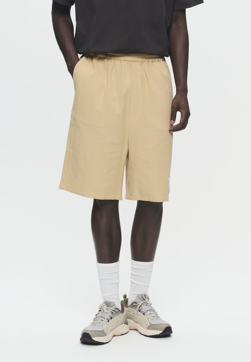 Beige shorts with an elastic waistband, made from textured fabric. Paired with gray sneakers and white crew socks.