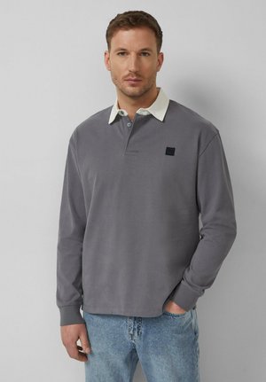 Homme portant un polo à manches longues gris avec un col blanc et un jean bleu clair, debout devant un fond uni gris clair.