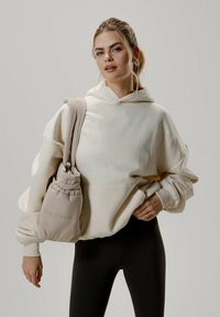 Sweat à capuche crème au fit oversized et à cordon de serrage, associé à un sac beige doux. Le mannequin porte des leggings noirs. Textures lisses partout.