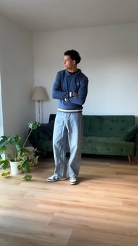 Dunkelblauer Hoodie mit langen Ärmeln, hellblaue lässige Jeans und graue Sneakers. Innenraum mit einem grünen Sofa und einer Topfpflanze. Natürliches Licht.