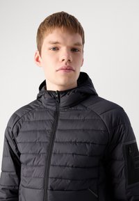 Casaco acolchoado preto com fecho de correr, capuz e padrões cosidos. Apresenta um bolso com fecho de correr na manga esquerda com uma marca.