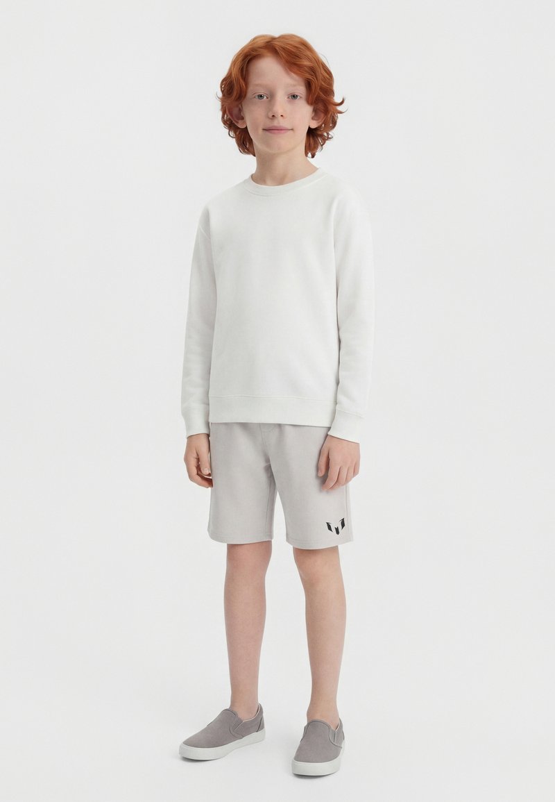 Ένα αγόρι με κόκκινα μαλλιά στέκεται προς τα εμπρός φορώντας ένα λευκό μακρυμάνικο πουκάμισο, ανοιχτό γκρι σορτς με λογότυπο και γκρι παπούτσια χωρίς κορδόνια σε λευκό φόντο.