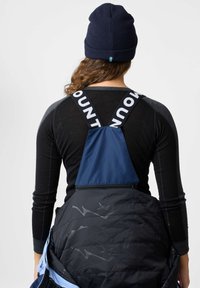 Schwarzes Langarmshirt mit grauen Akzenten, eine blaue wasserdichte Latzhose mit einem Logo-Träger und eine marineblaue Mütze mit einem Detail der schwedischen Flagge.