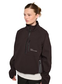 Femme à la peau claire et aux cheveux bruns en chignon portant une veste pull ample marron foncé avec des poches zippées et le texte « HALO 196819721981 ».