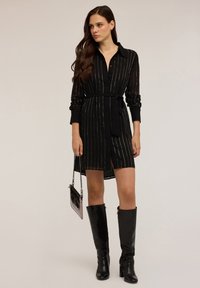 Abito camicia nero con strisce argentate verticali, colletto, maniche lunghe, vita cinturata e orlo a ginocchio. Indossato con stivali neri fino al ginocchio.