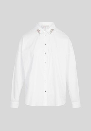 Camicia bianca a maniche lunghe con bottoni e decorazioni in argento e perle sul colletto, esposta su uno sfondo grigio uniforme.