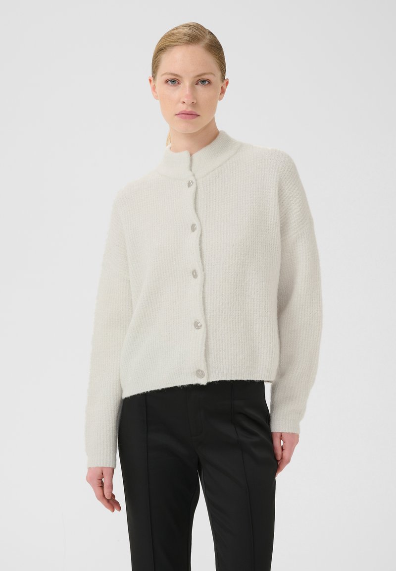 Gilet blanc court en tissu tricoté texturé, avec un col montant et de gros boutons. Associé à un pantalon noir ajusté.