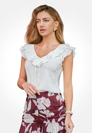 Frau mit welligem, hellbraunem Haar, die goldene Ohrringe, ein weißes ärmelloses Oberteil mit Rüschen und einen bordeauxroten Rock mit großem weißem Blumenmuster trägt.