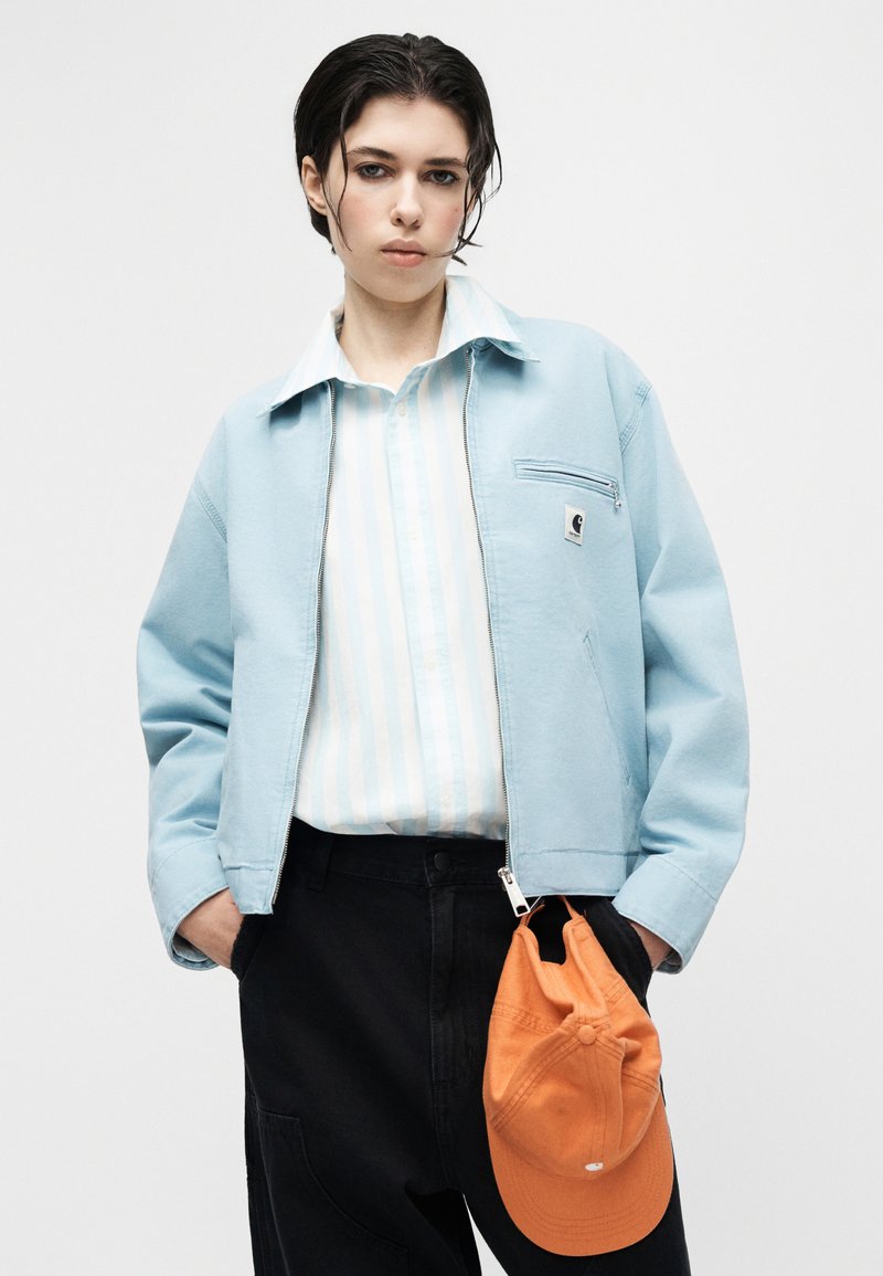 Personne aux cheveux courts et foncés portant une veste bleu clair par-dessus une chemise rayée, un pantalon noir, tenant une casquette de baseball orange dans la main droite.