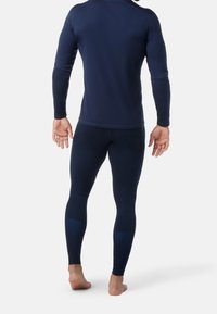 Man som bär en marinblå långärmad tröja och matchande tights, står barfota och vänder sig bort mot en vit bakgrund.