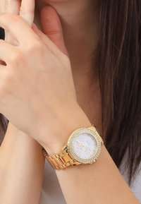 Kultakello, jossa on pyöreä kanta, valkoinen kellotaulu, kolme pienempää taulua ja kristallikoristeinen kehys. Varustettu kultasävyisellä metallirannekkeella.