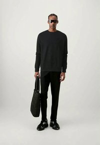 Top preto de mangas longas, calças pretas, ténis pretos com acentos brancos e uma mala tote preta com acolchoado. Modelo usa óculos de sol escuros.