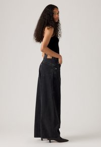 Czarne dżinsy o wysokim stanie i szerokich nogawkach, wykonane z denimu, z brązową skórzaną naszywką na plecach, noszone z czarnymi szpilkami o spiczastym nosku.