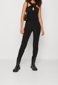 Zwart gerimpeld topje met bloemenkeyhole-detail, gecombineerd met strakke zwarte broeken en stevige zwarte enkelboots. Inclusief kleine zwarte crossbody tas.