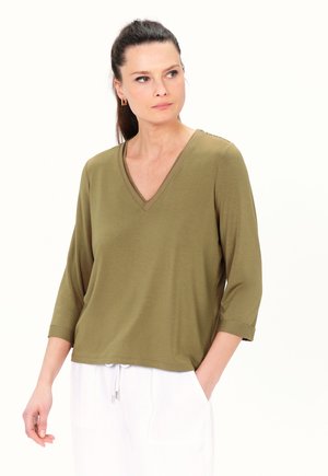 Olivgrünes V-Ausschnitt-Shirt mit Dreiviertelärmeln, glatter Textur und umgeschlagenen Bündchen. Kombination mit weißen Hosen mit Kordelzug.