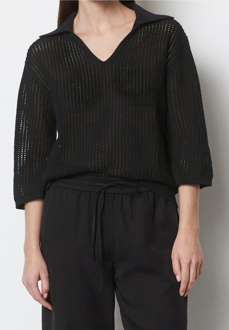 Femme portant un pull noir à maille lâche avec des manches trois-quarts et un pantalon noir à cordon, debout devant un fond clair uni.