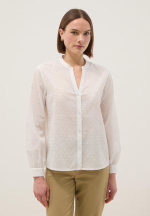 Donna che indossa una camicetta bianca trasparente a maniche lunghe con sottili pois e pantaloni beige, in piedi davanti a uno sfondo chiaro e uniforme.