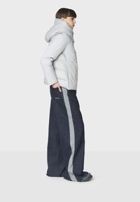 Giubbotto imbottito grigio chiaro con colletto alto e cappuccio, abbinato a jeans in denim scuro a gamba larga con una striscia grigia laterale.