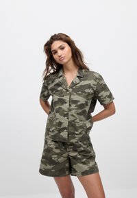 Camicia a maniche corte con motivo mimetico verde e marrone, abbinata a shorts coordinati. Il tessuto sembra leggero con una texture morbida.