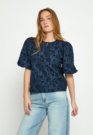 Žena s dlouhými blond vlasy, která má na sobě tmavě modrou květinovou blůzu s nabíranými rukávy a světlé vysoko pasové džíny, stojící na bílém pozadí.