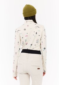 Dlhý rukáv krémového topu s farebnými abstraktnými vzormi, spojený s priliehavými svetlými nohavicami a zelenou pletenou čiapkou. Obsahuje čierny pás.