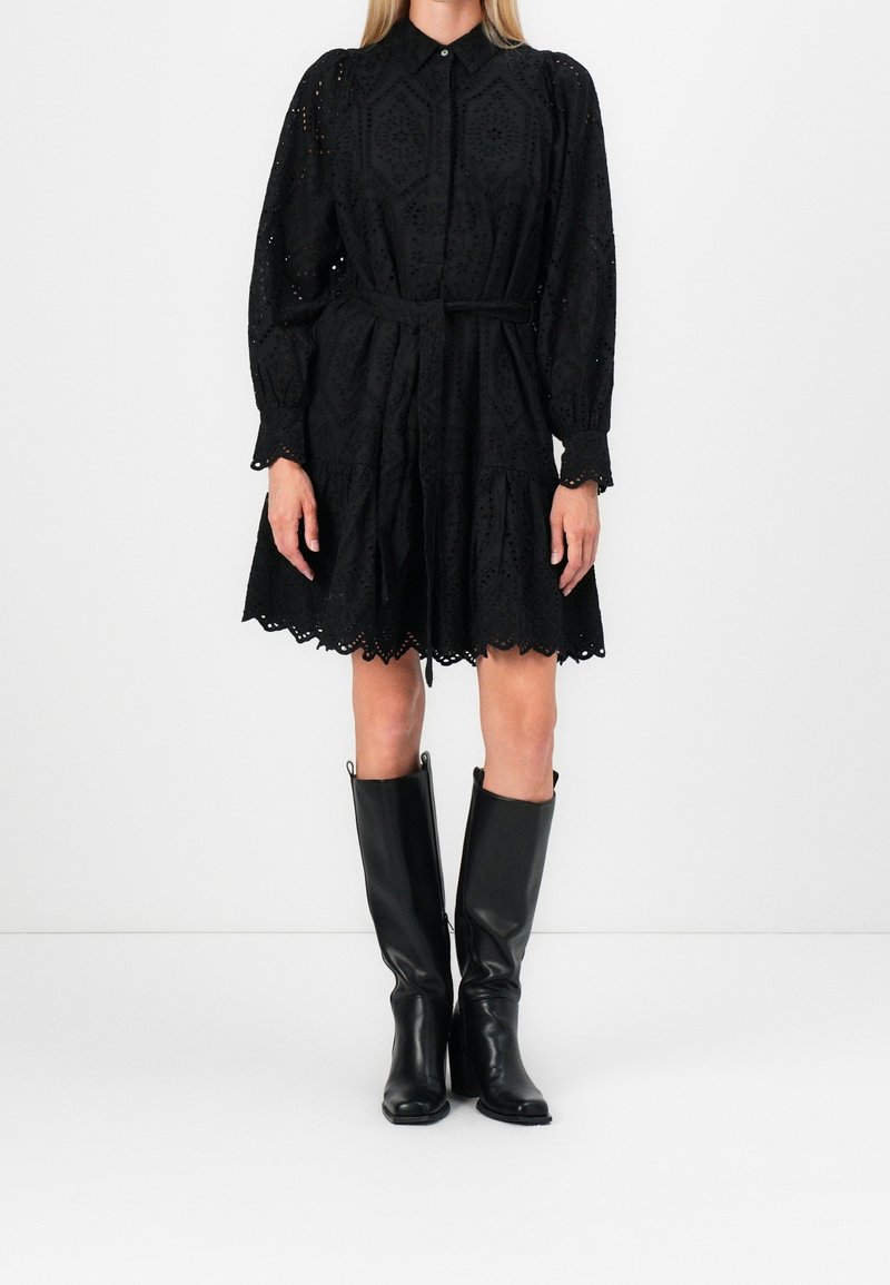 Femme portant une robe noire à manches longues avec des œillets et une ceinture, associée à des bottes en cuir noires remontant aux genoux, debout sur un fond blanc.