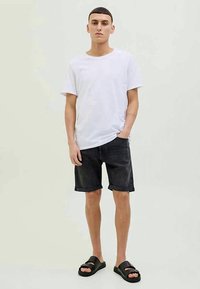 White cotton t-shirt, black denim shorts, and black slide sandals. Model stands confidently with hands in pockets, neutral background.
