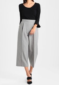 Schwarzes langärmliges Oberteil kombiniert mit hoch taillierten, weit geschnittenen grauen Wollhosen. Das Outfit besticht durch klare Linien und eine glatte Textur. Schwarze Absätze runden den Look ab.