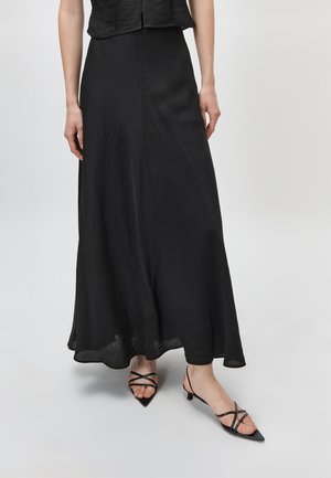 Žena nosi dlhú čiernu splývavú sukňu a čierne sandále s remienkami, špicatou špičkou a nízkym podpätkom, stojaca na svetlosivej podlahe.